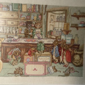 Peut inclure: Point de croix encadré représentant une scène de magasin avec des animaux anthropomorphes. Un chat et un chien sont derrière un comptoir, tandis que des lapins et des souris regardent. Le magasin est rempli d'étagères de bocaux et de boîtes, avec le texte "BREEDA TEA".
