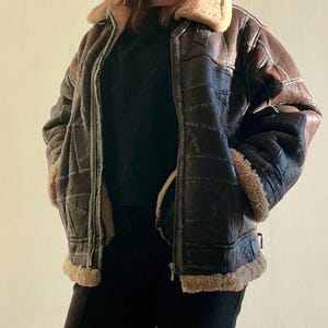 Könnte beinhalten: Braune Lederjacke mit Shearling-Kragen und -Besatz. Die Jacke hat ein Patchwork-Design und einen Reißverschluss. Innen und Kragen sind mit hellbraunem Shearling gefüttert. Die Jacke hat zwei Vordertaschen.