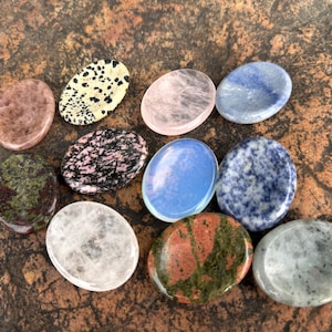 May include: A collection of oval-shaped polished gemstones in various colors and patterns. The stones include pink, blue, green, black and white, and speckled designs. The gemstones are displayed on a textured brown surface.