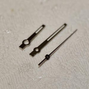May include: Close-up of three silver watch hands. The hands are different lengths and have circular or rectangular openings near the base. The longest hand is a thin, straight line with a small circle at the end. The hands are for watch repair or watchmaking.