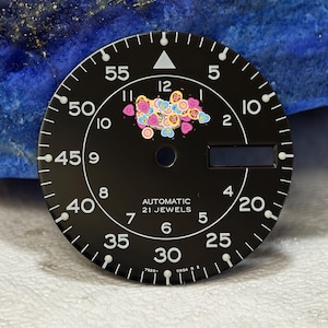 Könnte beinhalten: Schwarzes Zifferblatt mit weißen Ziffern und Zeigern, mit dem Text "AUTOMATIC 21 JEWELS". Verziert mit bunten herzförmigen Verzierungen. Das Zifferblatt hat einen Durchmesser von etwa 3,8 cm.