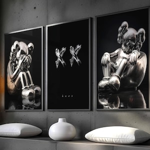 May include: A modern interior scene featuring three framed art prints with black backgrounds. Two prints showcase metallic sculptures, while the central print displays silver "XX" symbols. The room includes a concrete ledge with white pillows and a vase, and a hanging lamp.