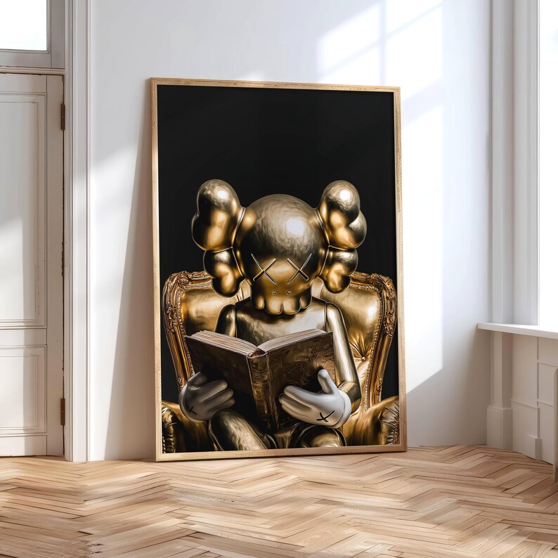 Puede incluir: Una impresi&oacute;n art&iacute;stica enmarcada que presenta una escultura dorada de una figura con los ojos tachados, sentada en una silla dorada ornamentada, leyendo un libro. El fondo es negro y el marco es dorado. La obra de arte se exhibe contra una pared blanca.