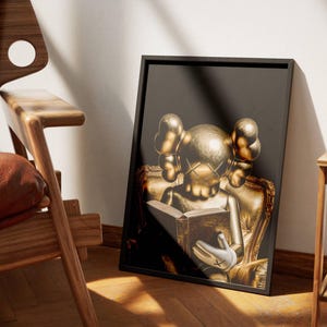 Puede incluir: Una impresi&oacute;n art&iacute;stica enmarcada que presenta una figura dorada leyendo un libro, sobre un fondo oscuro. La figura est&aacute; sentada en una silla dorada. La obra de arte se muestra en un marco negro, apoyada contra una pared cerca de una silla de madera.