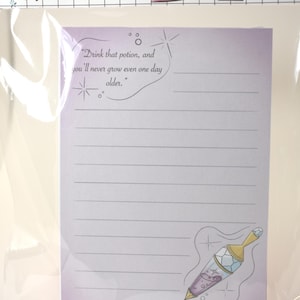 May include: A lined notepad with a light purple background, featuring a potion illustration and the text "Drink that potion, and you'll never grow even one day older." The notepad is packaged in clear plastic.
