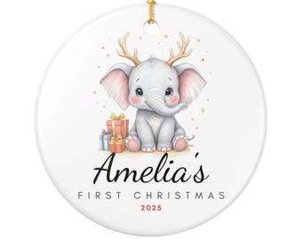May include: White ceramic Christmas ornament featuring a baby elephant with antlers, sitting next to wrapped gifts. The ornament has the text "Amelia's FIRST CHRISTMAS 2025" in black and red. A gold string is attached for hanging.