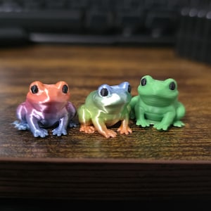 Puede incluir: Tres pequeñas figuras de ranas coloridas. Una es un degradado de rojo, morado y azul. La segunda es verde, amarillo y naranja. La tercera es verde liso. Todas tienen ojos negros y están sobre una superficie de madera.