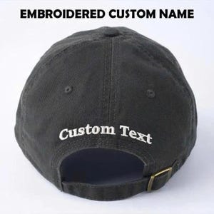 May include: A dark gray baseball cap with the text "EMBROIDERED CUSTOM NAME" at the top and "Custom Text" embroidered on the back. The cap has a curved brim and a metal clasp.
