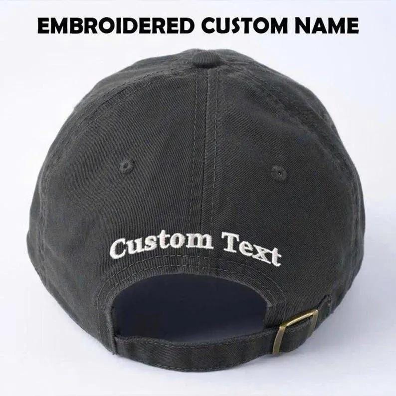 May include: A dark gray baseball cap with the text "EMBROIDERED CUSTOM NAME" at the top and "Custom Text" embroidered on the back. The cap has a curved brim and a metal clasp.