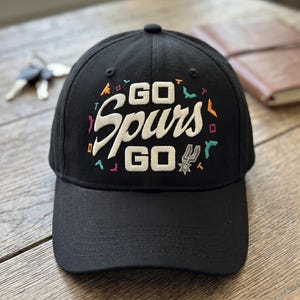 May include: Black baseball cap with the words "GO Spurs GO" embroidered in white script. The cap features colorful accents and the Spurs logo. A stylish accessory for fans.