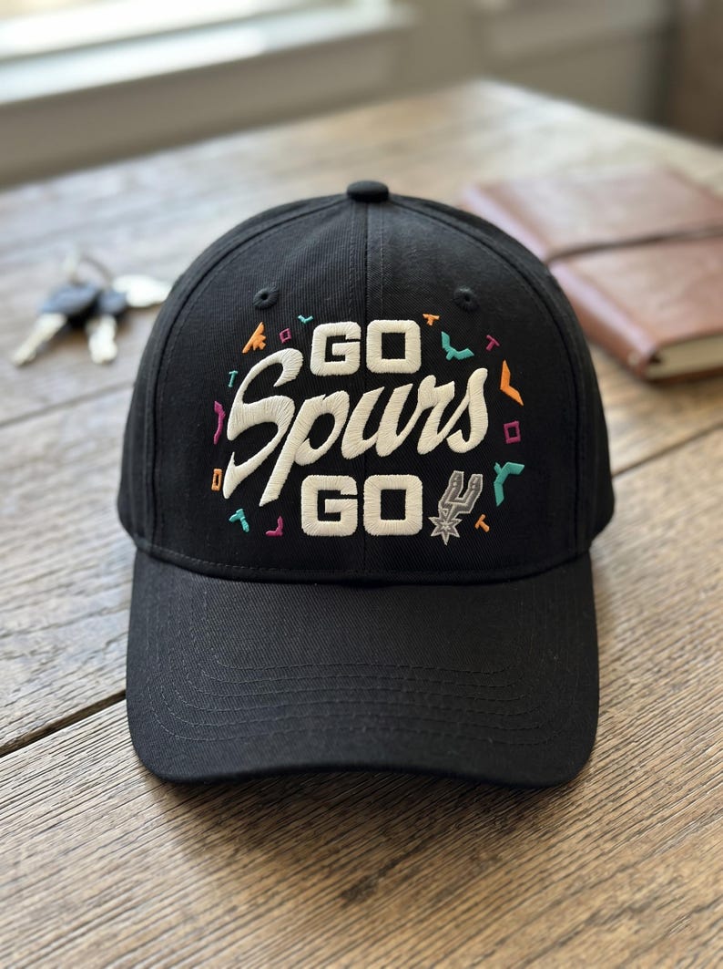 May include: Black baseball cap with the words "GO Spurs GO" embroidered in white script. The cap features colorful accents and the Spurs logo. A stylish accessory for fans.