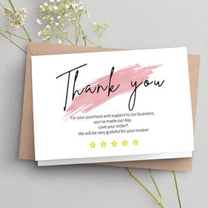 May include: A white card with the words "Thank you" in black script, over a pink brushstroke. The card includes text expressing gratitude and a row of five gold stars. The card is placed on a brown envelope.