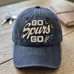 May include: A navy blue baseball cap with the embroidered text "GO Spurs GO" in white, with colorful accents. The cap features the Spurs logo and is sitting on a wooden surface.
