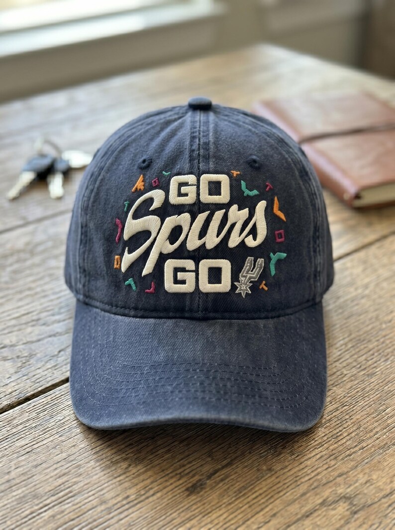 May include: A navy blue baseball cap with the embroidered text "GO Spurs GO" in white, with colorful accents. The cap features the Spurs logo and is sitting on a wooden surface.