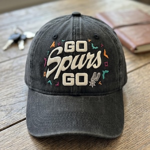 May include: A black baseball cap with the text "GO Spurs GO" embroidered in white, with colorful accents. The cap features the Spurs logo and is made of a worn, faded fabric. A notebook and keys are in the background.