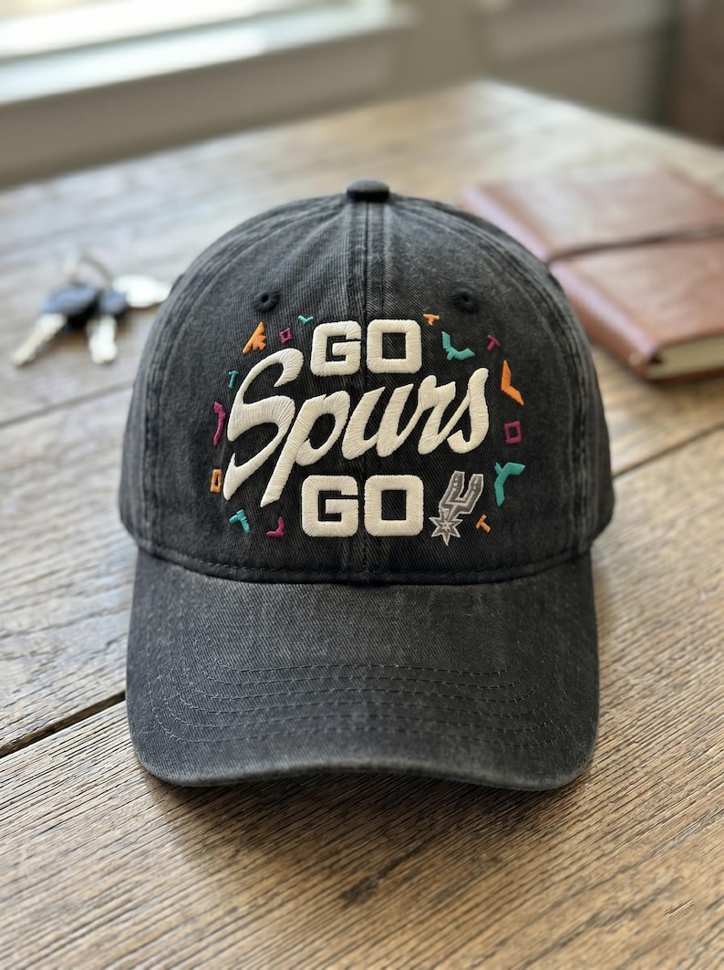 May include: A black baseball cap with the text "GO Spurs GO" embroidered in white, with colorful accents. The cap features the Spurs logo and is made of a worn, faded fabric. A notebook and keys are in the background.