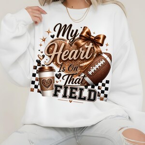Puede incluir: Una sudadera blanca de manga larga con un estampado gr&aacute;fico que incluye el texto My Heart Is On That FIELD. El dise&ntilde;o presenta un coraz&oacute;n dorado brillante, un bal&oacute;n de f&uacute;tbol americano, una taza de caf&eacute; con un coraz&oacute;n de leopardo, un lazo marr&oacute;n y cuadros negros.