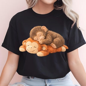 May include: Black t-shirt featuring a graphic of two sleeping monkeys. One monkey is brown and resting on top of a larger orange monkey. The image has a soft, cartoon-like style.