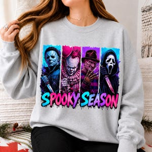 May include: A light gray crewneck sweatshirt featuring a graphic print of four horror movie characters in vertical panels. Below the characters, the text SPOOKY SEASON is printed in a vibrant blue and pink gradient with a dripping paint effect.