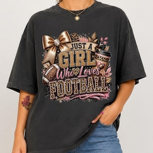 Puede incluir: Una camiseta gris carb&oacute;n con un estampado gr&aacute;fico que incluye el texto JUST A GIRL Who Loves FOOTBALL. El dise&ntilde;o presenta un lazo dorado grande, un vaso de caf&eacute; con estampado de leopardo, un bal&oacute;n de f&uacute;tbol americano y detalles en purpurina rosa.