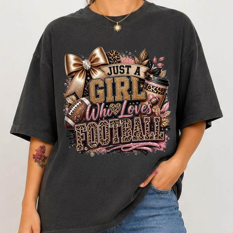 Puede incluir: Una camiseta gris carb&oacute;n con un estampado gr&aacute;fico que incluye el texto JUST A GIRL Who Loves FOOTBALL. El dise&ntilde;o presenta un lazo dorado grande, un vaso de caf&eacute; con estampado de leopardo, un bal&oacute;n de f&uacute;tbol americano y detalles en purpurina rosa.