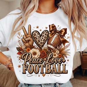 Puede incluir: Una sudadera blanca con un estampado gr&aacute;fico que incluye un signo de paz con purpurina dorada, un coraz&oacute;n con estampado de leopardo y un bal&oacute;n de f&uacute;tbol americano con un gran lazo marr&oacute;n. El texto Peace Love FOOTBALL en oro rodeado de flores.