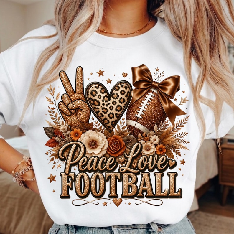Puede incluir: Una sudadera blanca con un estampado gr&aacute;fico que incluye un signo de paz con purpurina dorada, un coraz&oacute;n con estampado de leopardo y un bal&oacute;n de f&uacute;tbol americano con un gran lazo marr&oacute;n. El texto Peace Love FOOTBALL en oro rodeado de flores.
