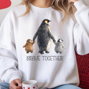 May include: White sweatshirt featuring a watercolor illustration of a penguin family. The adult penguin holds the hands of two smaller penguins. The text "BRAVE TOGETHER" is printed below the illustration. The sweatshirt is a comfortable, casual style.