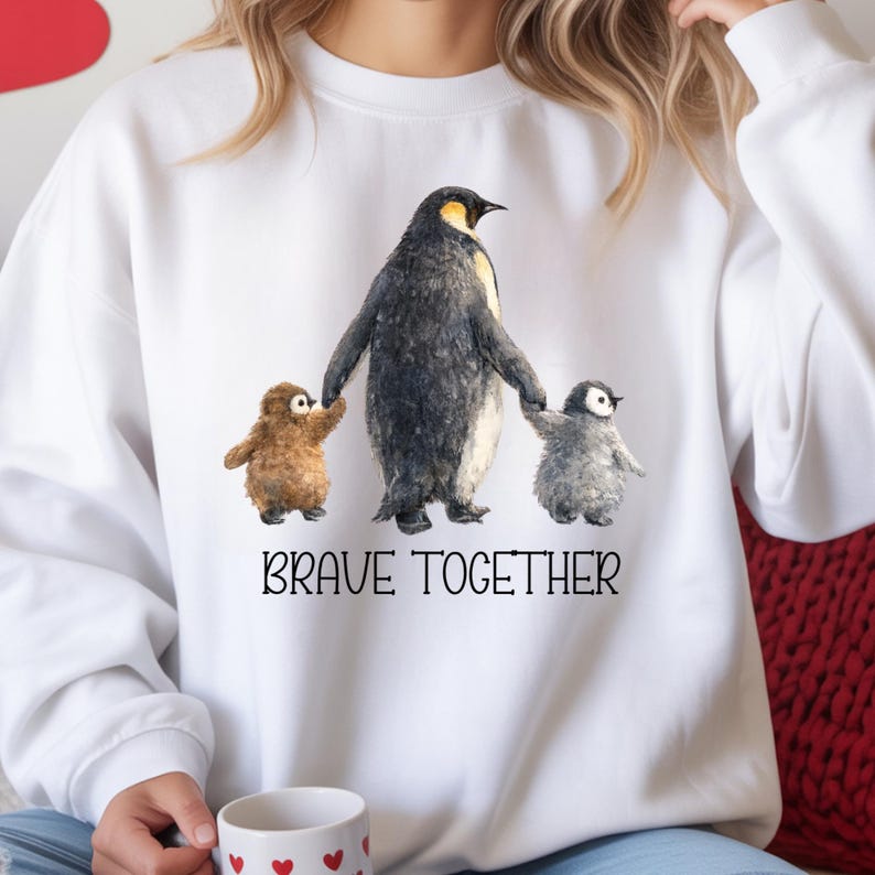 May include: White sweatshirt featuring a watercolor illustration of a penguin family. The adult penguin holds the hands of two smaller penguins. The text "BRAVE TOGETHER" is printed below the illustration. The sweatshirt is a comfortable, casual style.