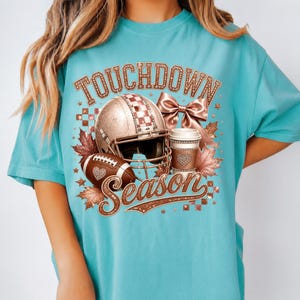 Puede incluir: Una camiseta turquesa con un estampado gr&aacute;fico que incluye el texto TOUCHDOWN SEASON. El dise&ntilde;o presenta un casco de f&uacute;tbol americano en oro rosa, un bal&oacute;n, una taza de caf&eacute;, un lazo y hojas de oto&ntilde;o, con texturas y patrones brillantes.