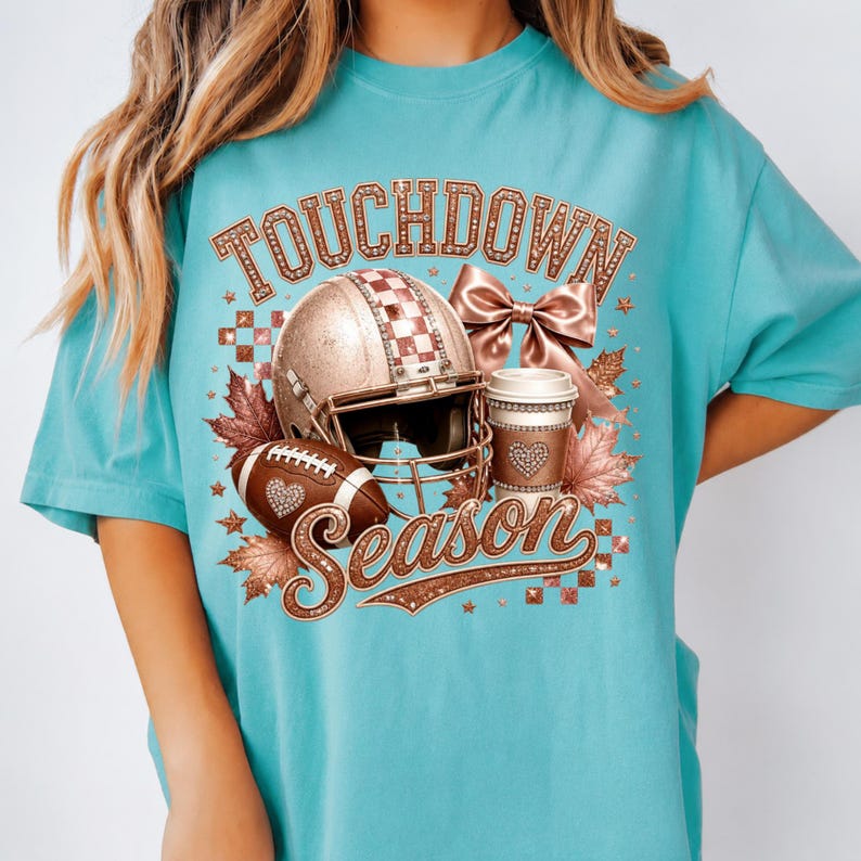 Puede incluir: Una camiseta turquesa con un estampado gr&aacute;fico que incluye el texto TOUCHDOWN SEASON. El dise&ntilde;o presenta un casco de f&uacute;tbol americano en oro rosa, un bal&oacute;n, una taza de caf&eacute;, un lazo y hojas de oto&ntilde;o, con texturas y patrones brillantes.