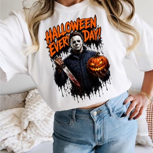 May include: A white t-shirt featuring a graphic print of a masked figure in a dark jacket holding a bloody knife and a glowing jack-o'-lantern. The text HALLOWEEN EVERY DAY is printed in orange above the figure, with black ink drip effects.