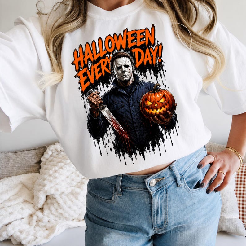 May include: A white t-shirt featuring a graphic print of a masked figure in a dark jacket holding a bloody knife and a glowing jack-o'-lantern. The text HALLOWEEN EVERY DAY is printed in orange above the figure, with black ink drip effects.