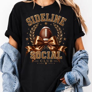 Puede incluir: Una camiseta negra con un dise&ntilde;o gr&aacute;fico que incluye el texto SIDELINE SOCIAL CLUB en letras doradas con purpurina. El dise&ntilde;o presenta un bal&oacute;n de f&uacute;tbol americano marr&oacute;n, un gran lazo dorado met&aacute;lico, ramas de laurel y estrellas doradas.