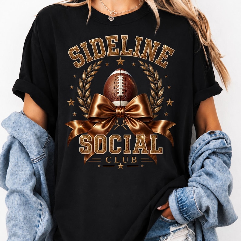 Puede incluir: Una camiseta negra con un dise&ntilde;o gr&aacute;fico que incluye el texto SIDELINE SOCIAL CLUB en letras doradas con purpurina. El dise&ntilde;o presenta un bal&oacute;n de f&uacute;tbol americano marr&oacute;n, un gran lazo dorado met&aacute;lico, ramas de laurel y estrellas doradas.