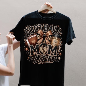 Puede incluir: Una camiseta negra de manga corta con un estampado gr&aacute;fico que incluye el texto FOOTBALL MOM LIFE. El dise&ntilde;o presenta un bal&oacute;n de f&uacute;tbol americano marr&oacute;n, un lazo con estampado de leopardo y un vaso de caf&eacute; con la frase Fueled by COFFEE TEAM SPIRIT.