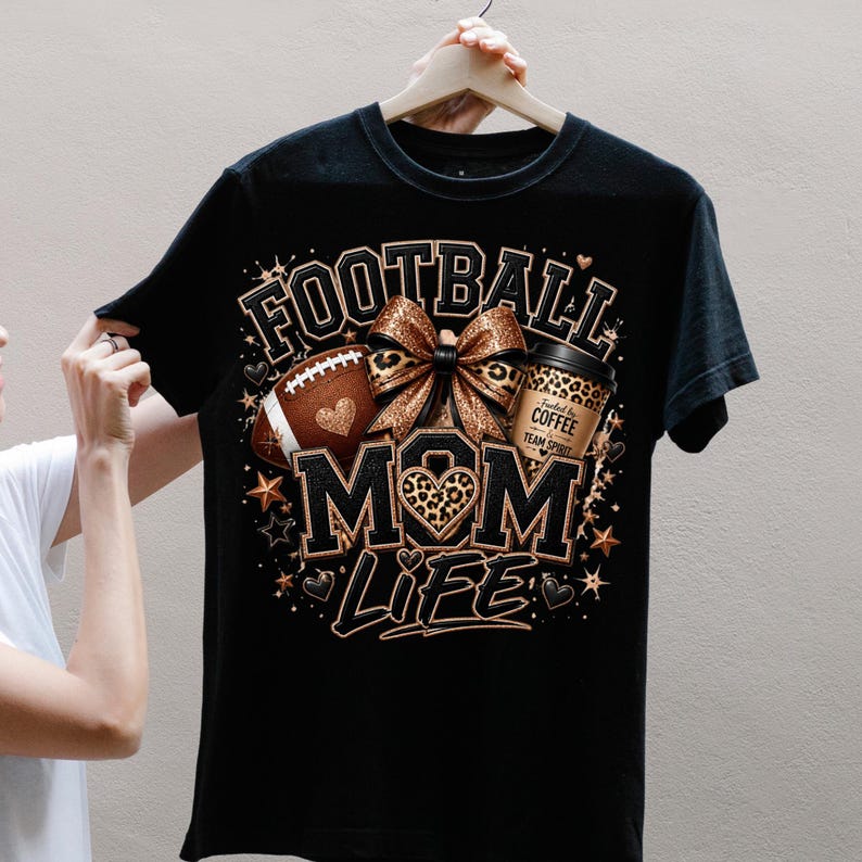 Puede incluir: Una camiseta negra de manga corta con un estampado gr&aacute;fico que incluye el texto FOOTBALL MOM LIFE. El dise&ntilde;o presenta un bal&oacute;n de f&uacute;tbol americano marr&oacute;n, un lazo con estampado de leopardo y un vaso de caf&eacute; con la frase Fueled by COFFEE TEAM SPIRIT.