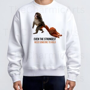 May include: White sweatshirt featuring a graphic of a monkey holding a stuffed animal. The text "EVEN THE STRONGEST NEED SOMEONE TO HOLD" is printed below the image in orange and black. The sweatshirt is a casual style.