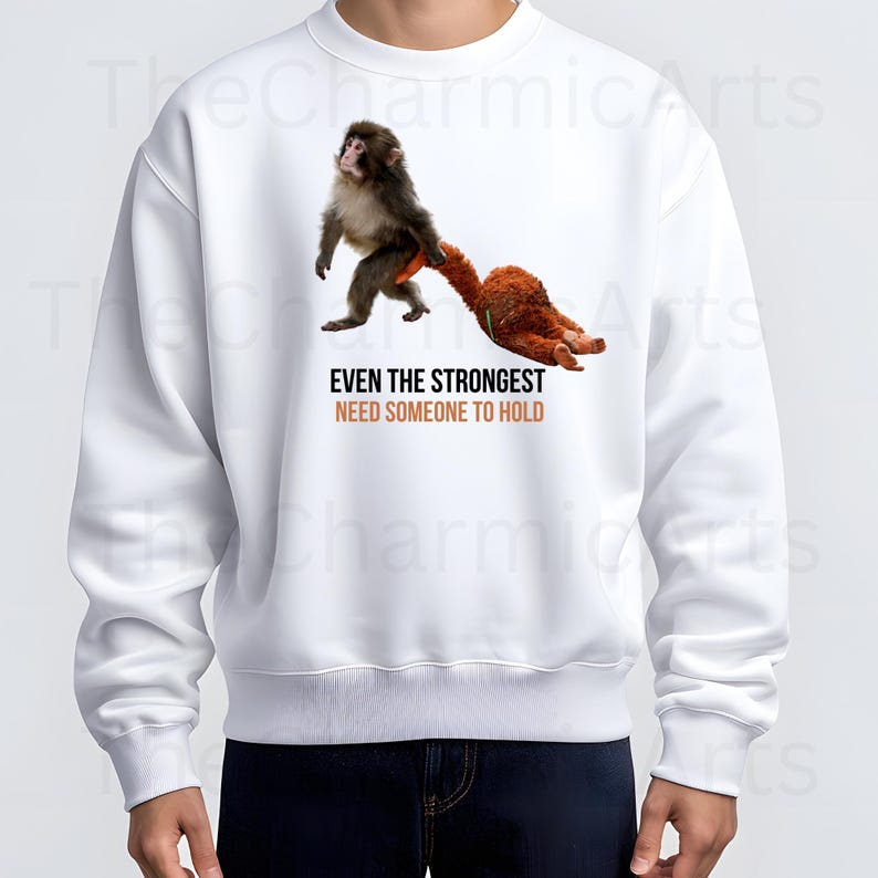 May include: White sweatshirt featuring a graphic of a monkey holding a stuffed animal. The text "EVEN THE STRONGEST NEED SOMEONE TO HOLD" is printed below the image in orange and black. The sweatshirt is a casual style.