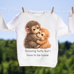 May include: White sweatshirt featuring a watercolor illustration of two hugging monkeys in shades of brown and orange. The text "Growing hurts, but I have to be brave" is printed below the image. The sweatshirt is hanging on a clothesline.