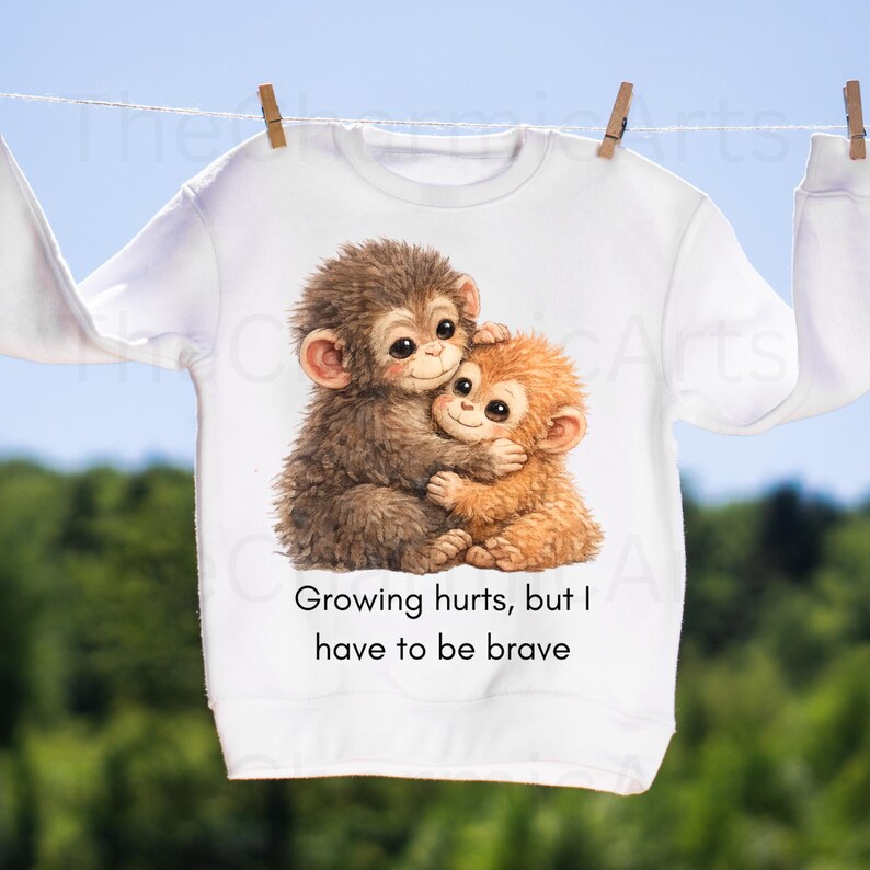 May include: White sweatshirt featuring a watercolor illustration of two hugging monkeys in shades of brown and orange. The text "Growing hurts, but I have to be brave" is printed below the image. The sweatshirt is hanging on a clothesline.