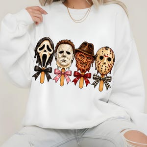 May include: A white long-sleeve sweatshirt featuring a graphic print of four horror movie character masks styled as popsicles on wooden sticks. Each popsicle is adorned with a decorative bow in black, pink, red, or black and white plaid patterns.