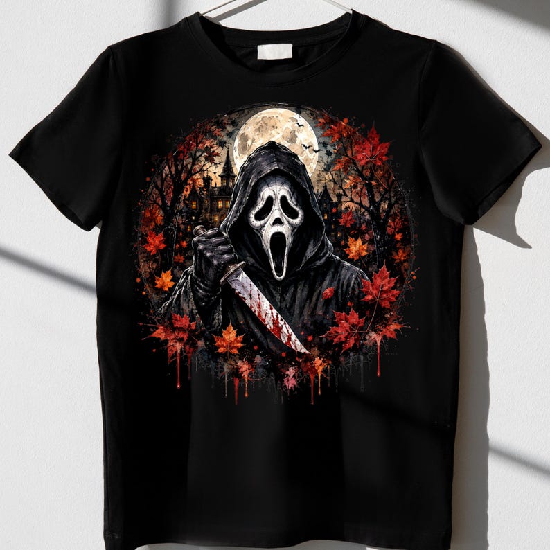May include: A black short-sleeve t-shirt featuring a graphic print of a hooded figure in a white mask holding a bloody knife. The design includes a full moon, a dark silhouette of a house, and vibrant red and orange autumn maple leaves.