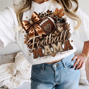 Puede incluir: Una camiseta blanca con un estampado gr&aacute;fico que incluye un bal&oacute;n de f&uacute;tbol americano marr&oacute;n, un lazo de sat&eacute;n, una taza de caf&eacute; con estampado de leopardo y hojas de oto&ntilde;o. El texto "Football MAMA" aparece en una fuente marr&oacute;n brillante.