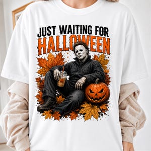 May include: A white t-shirt featuring a graphic of a character in a dark jumpsuit sitting among orange autumn leaves. The character holds a clear plastic cup with a pumpkin design. The text reads JUST WAITING FOR HALLOWEEN in black and orange.