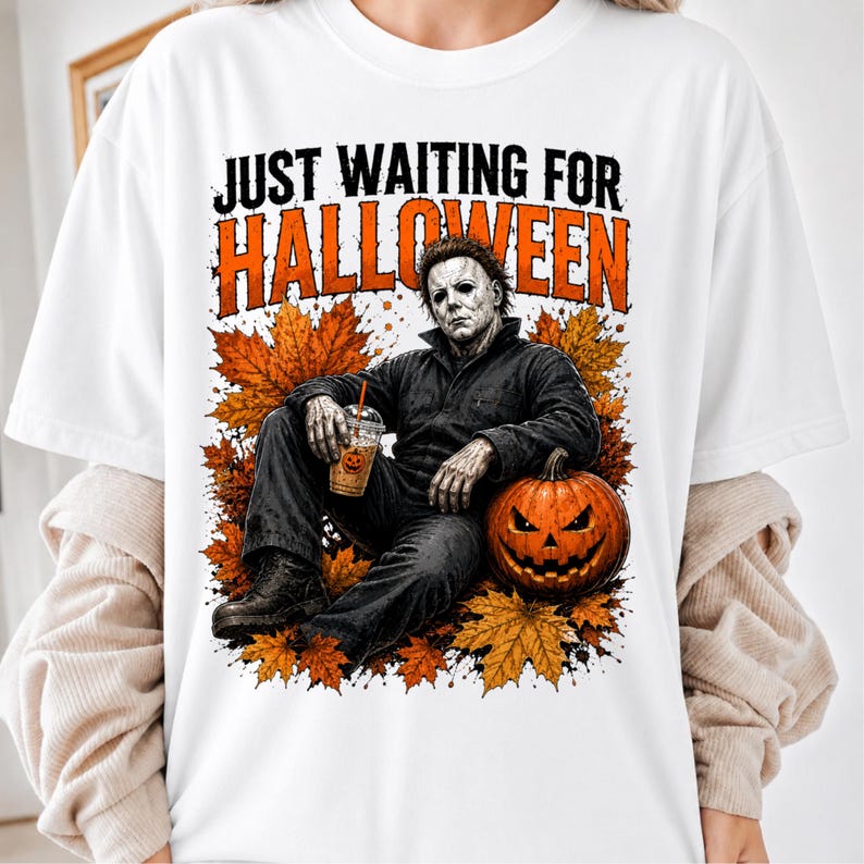May include: A white t-shirt featuring a graphic of a character in a dark jumpsuit sitting among orange autumn leaves. The character holds a clear plastic cup with a pumpkin design. The text reads JUST WAITING FOR HALLOWEEN in black and orange.