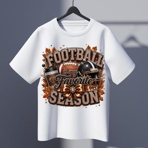 Puede incluir: Una camiseta blanca con un estampado gr&aacute;fico que incluye el texto "FOOTBALL is my FAVORITE SEASON". El dise&ntilde;o muestra un bal&oacute;n de f&uacute;tbol americano marr&oacute;n, un casco negro, una taza de caf&eacute; y un gran lazo marr&oacute;n, rodeados de hojas oto&ntilde;ales naranjas.