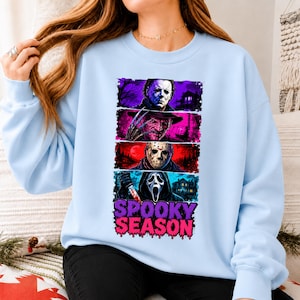 May include: A light blue crewneck sweatshirt featuring a graphic print of four horror movie characters stacked vertically in colorful panels. Below the characters, the text SPOOKY SEASON is printed in a bold, dripping purple and pink font.