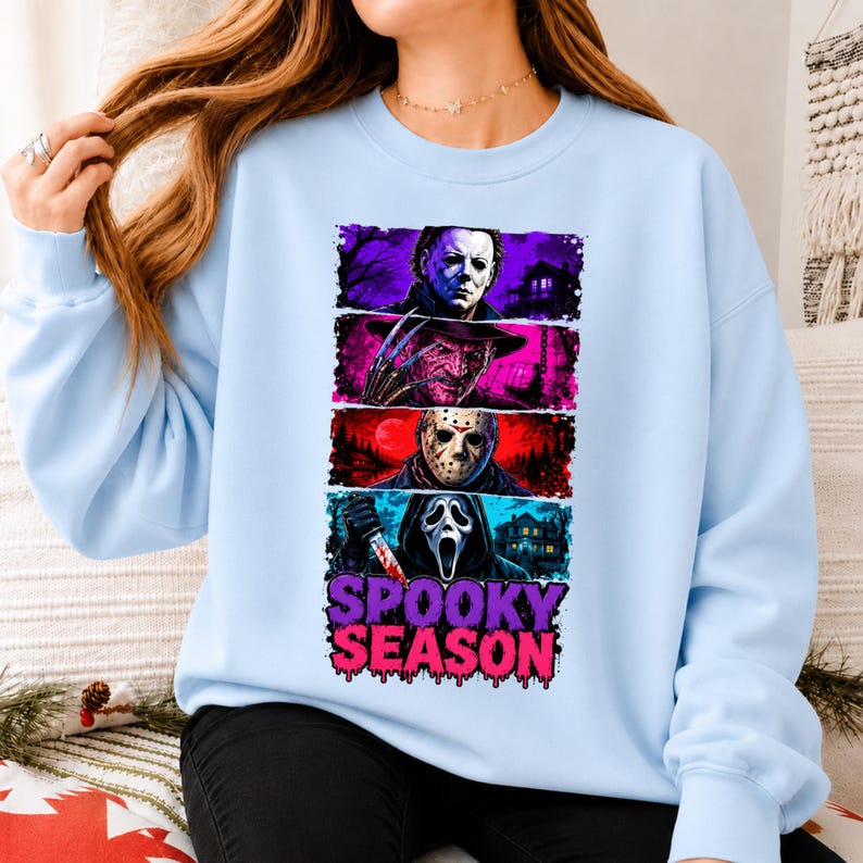 May include: A light blue crewneck sweatshirt featuring a graphic print of four horror movie characters stacked vertically in colorful panels. Below the characters, the text SPOOKY SEASON is printed in a bold, dripping purple and pink font.