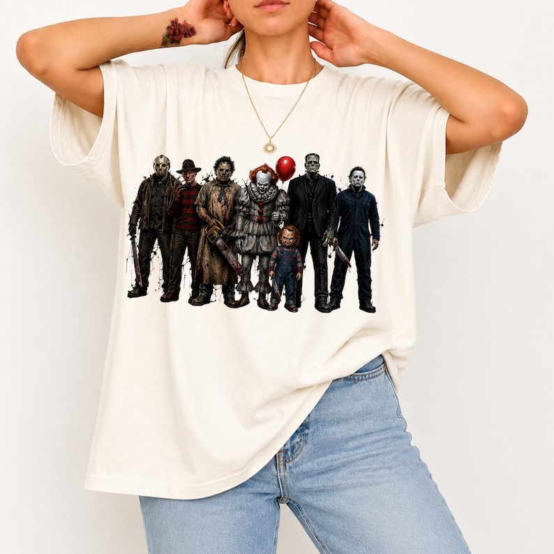 May include: An off-white short-sleeve t-shirt featuring a graphic print of seven iconic horror movie characters standing in a row. The design includes figures such as a clown with a red balloon, a doll, and various masked or costumed villains.